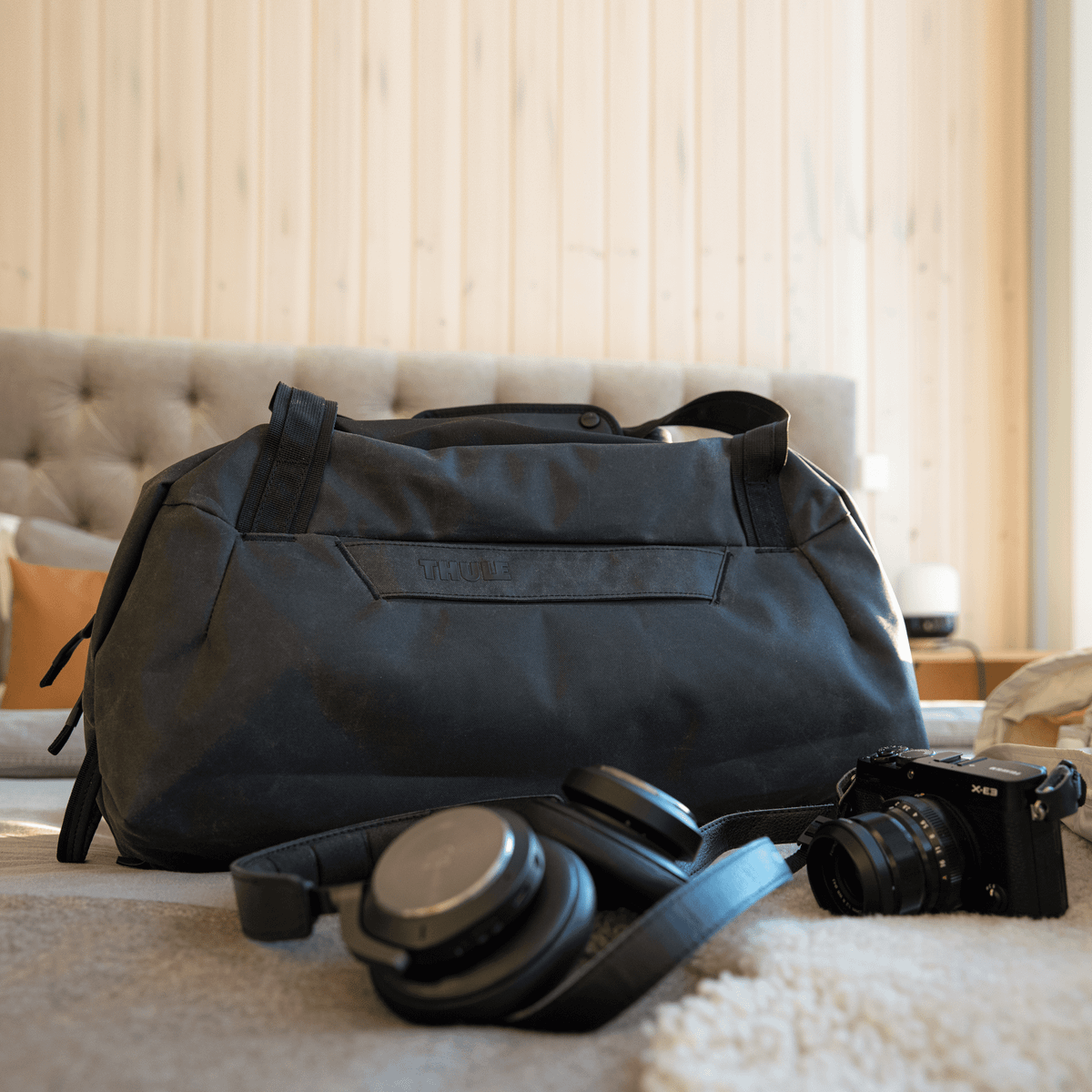 A black Thule Aion Duffel sits on a bed with headphones and a camera in a hotel room.