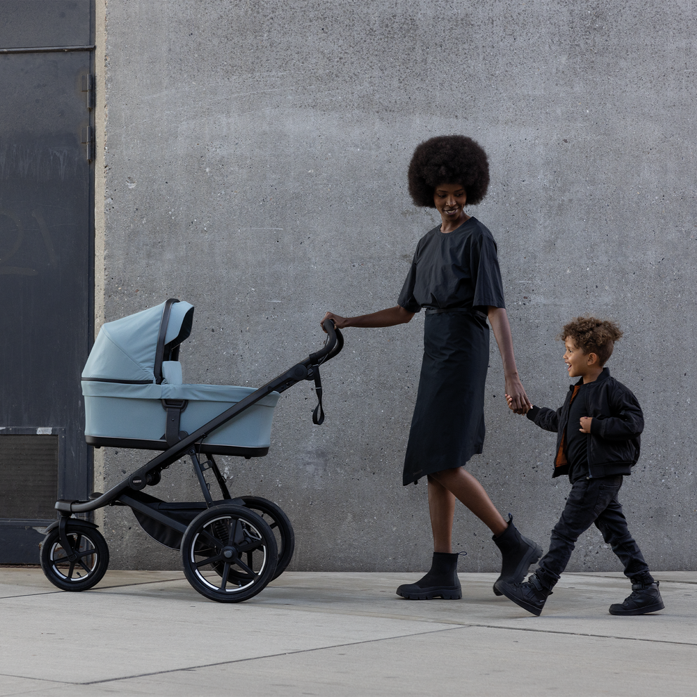 A mother walks with her son and her baby inside a blue Thule Urban Glide 3 bassinet stroller.