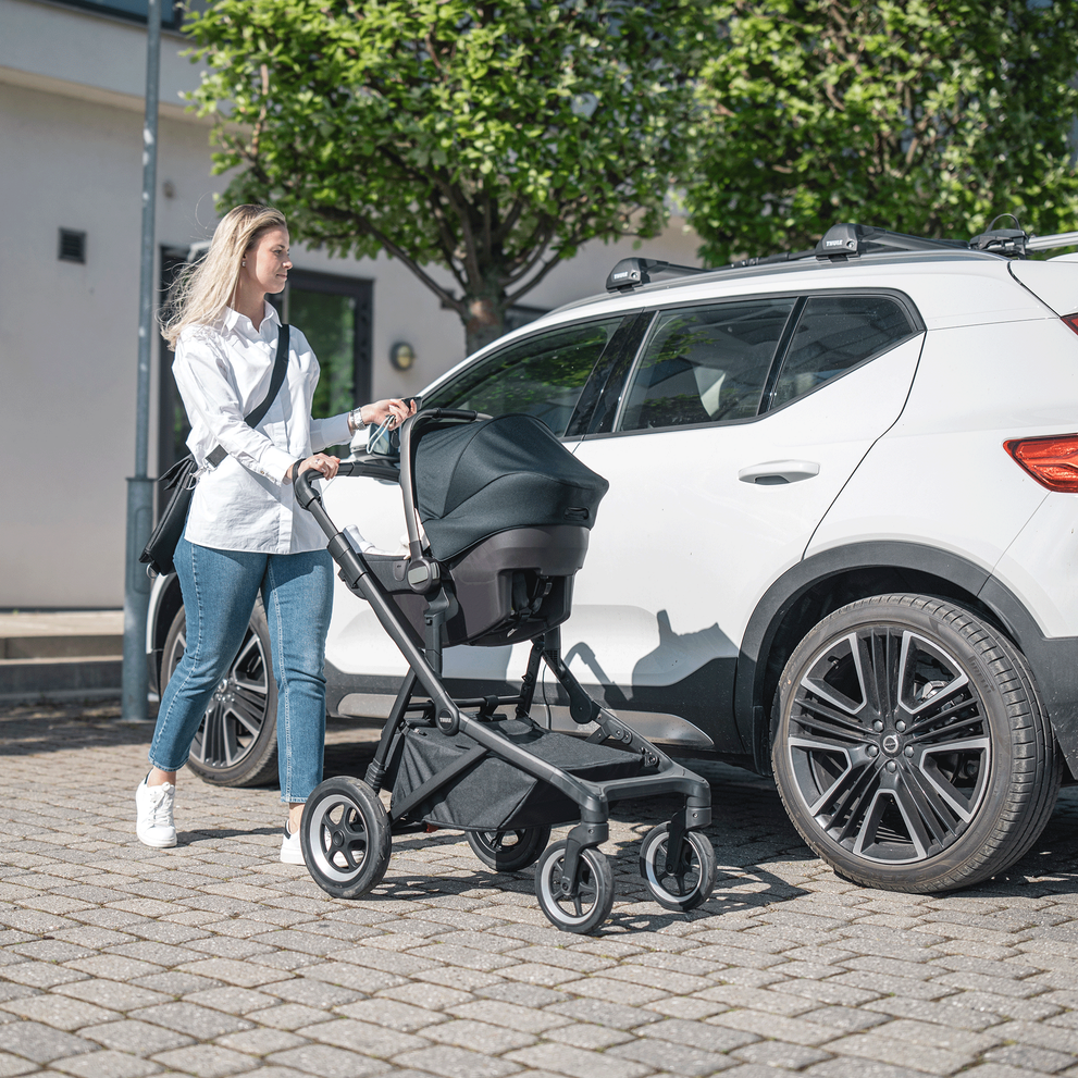 A woman walks away from her white car with her baby in a Thule Sleek baby stroller with car seat installed.