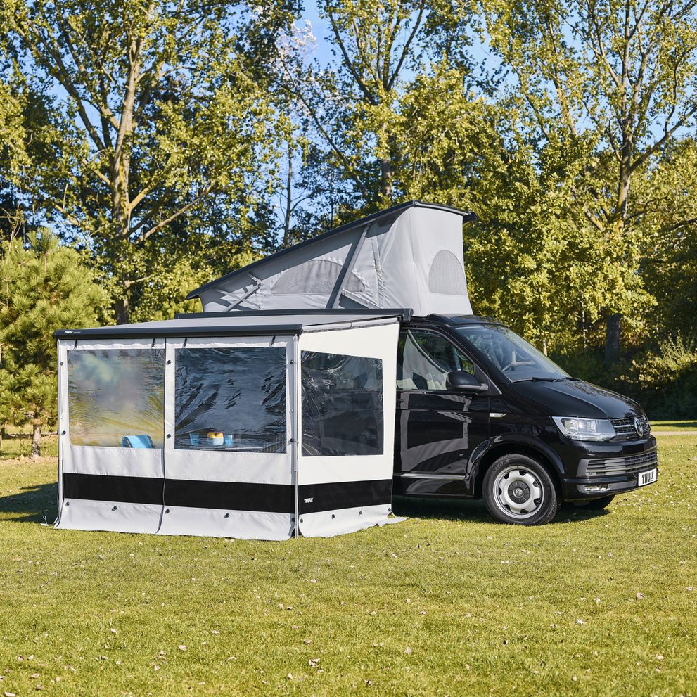 Compact van with pop-up roof and open Thule awning with panel set set up on a grass field in the sun.