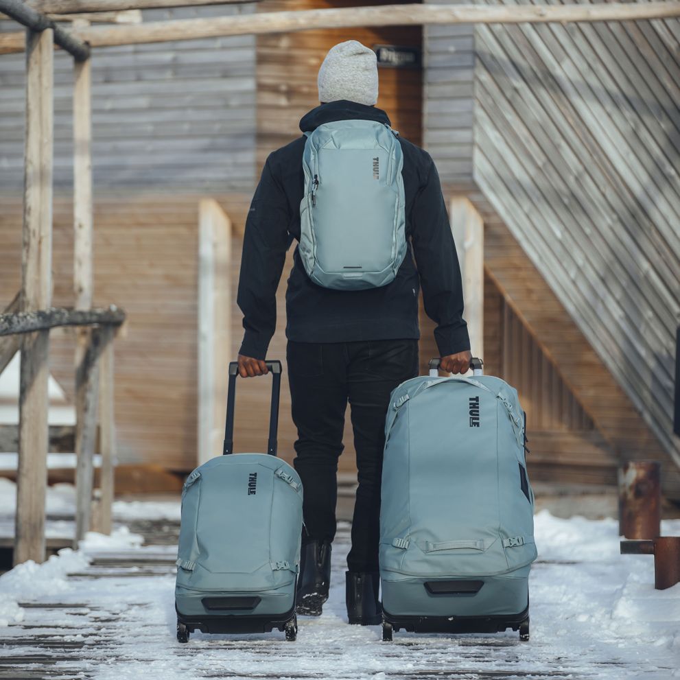 A man is walking down a snowy path with blue Thule Chasm suitcases and a backpack.