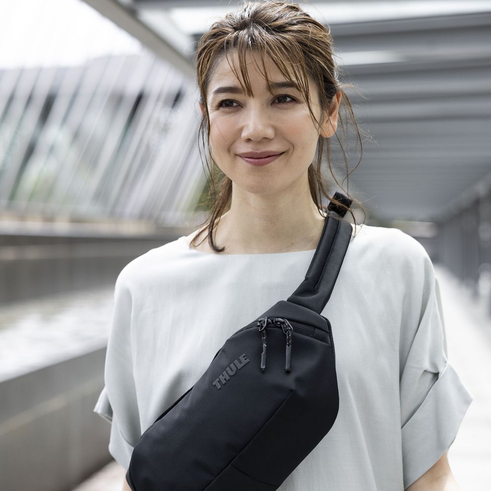 A woman in white looks off in the distance wearing a black Thule Subterra sling bag over her shoulder.