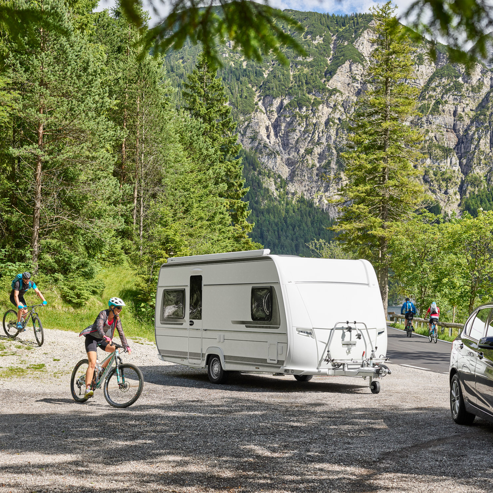 Two cyclists cycle down a forest path beside a caravan with a Thule Caravan Smart caravan bike rack.