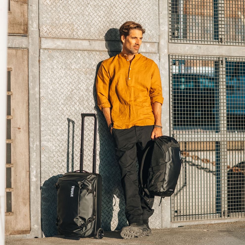 A man is leaning against a concrete wall holding a black Thule Chasm backpack next to a suitcase.