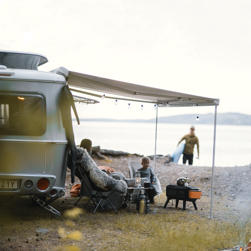 A man with a surfboard walks towards a woman sitting by a white van with a Thule Omnistor 6300 van awning.
