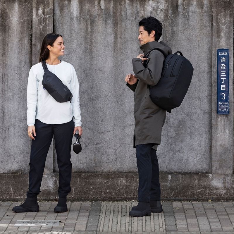 A man and woman stand next to a concrete wall, talking and carrying a Thule Subterra backpack and crossbody.