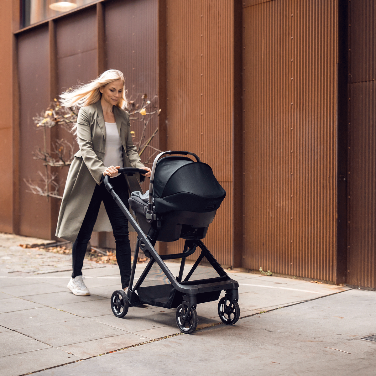 A woman walks down a rustic city street with her baby in a Thule Shine compact stroller with a car seat.
