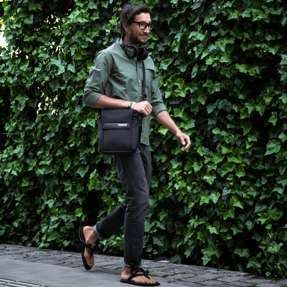 A man walks down the street with headphones, a green shirt and a Thule Paramount Crossbody Tote.