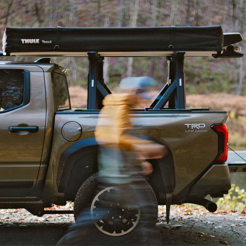 A man walks beside a vehicle with Thule Xscape truck rack that carries the rooftop tent Thule Foothill and the fishing rod rack Thule RodVault 2 in an outdoors environment.