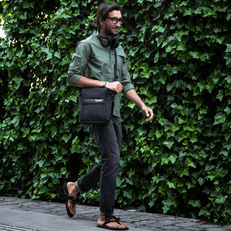A man walks down the street with headphones, a green shirt and a Thule Paramount crossbody tote.