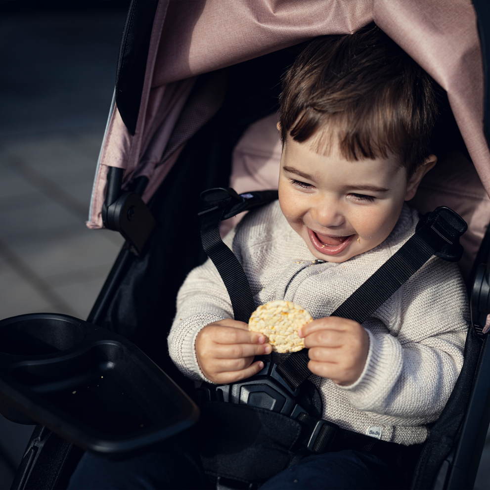 A toddler laughs holding a snack, sitting in a pink stroller with a Thule Spring Snack Tray.
