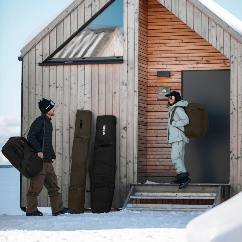 A man and a woman are standing outside a wooden house with their ski gear in Thule RoundTrip bags.