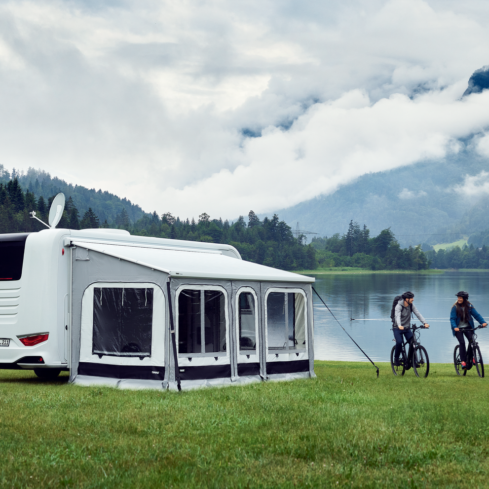 A motorhome with a Thule Panorama rv tent is parked by a lake in the moutains, and two people bike beside it.