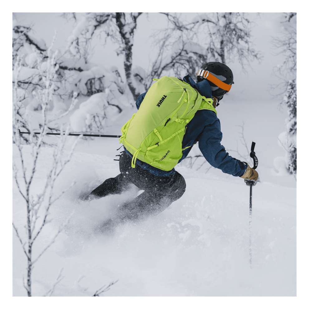 A skier skis down a snowy slope with a green Thule Upslope ski backpack.