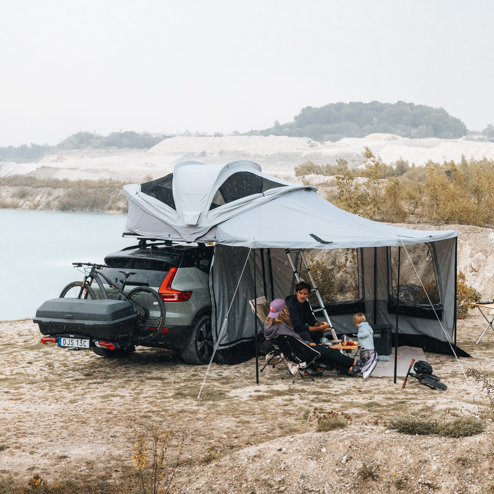 Thule Approach 2 soft-shell rooftop tent unfolded and mounted with awning and walls on the top of a car with one bike and Thule Santu cargo box on the towbar bike rack parked in an outdoor setting and a family interacting by the tent.