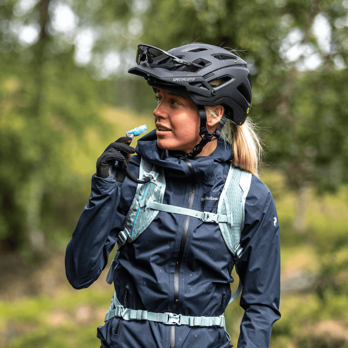 A woman on an mtb path with a black helmet takes a sip of water from a Thule Vital 3L Hydration Backpack.
