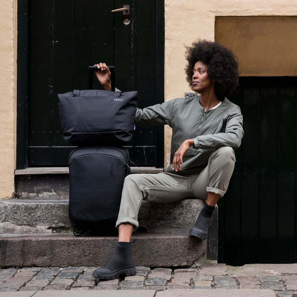A woman sits on a stoop holding a black Thule Subterra tote on the Thule subterra carry-on suitcase.