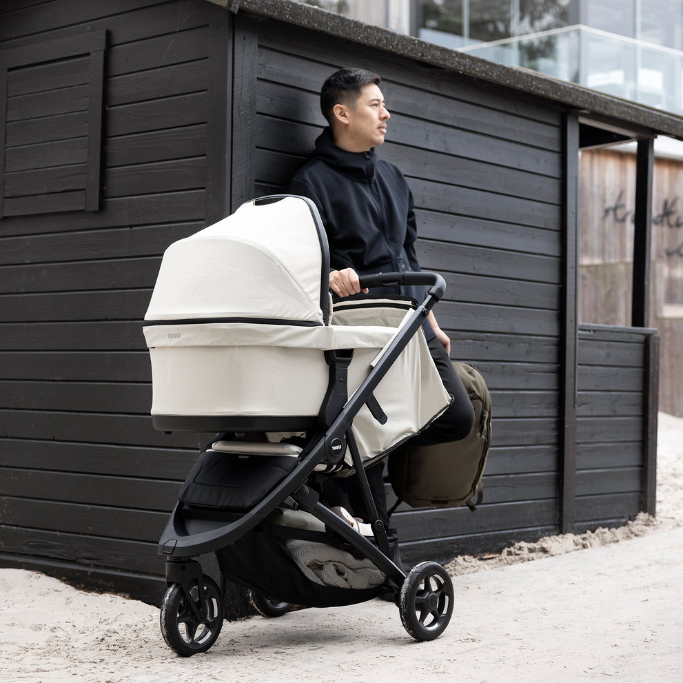 A man holding the bar of the Thule Spring 2 stroller with a light beige bassinet in front of a black wooden cabin.