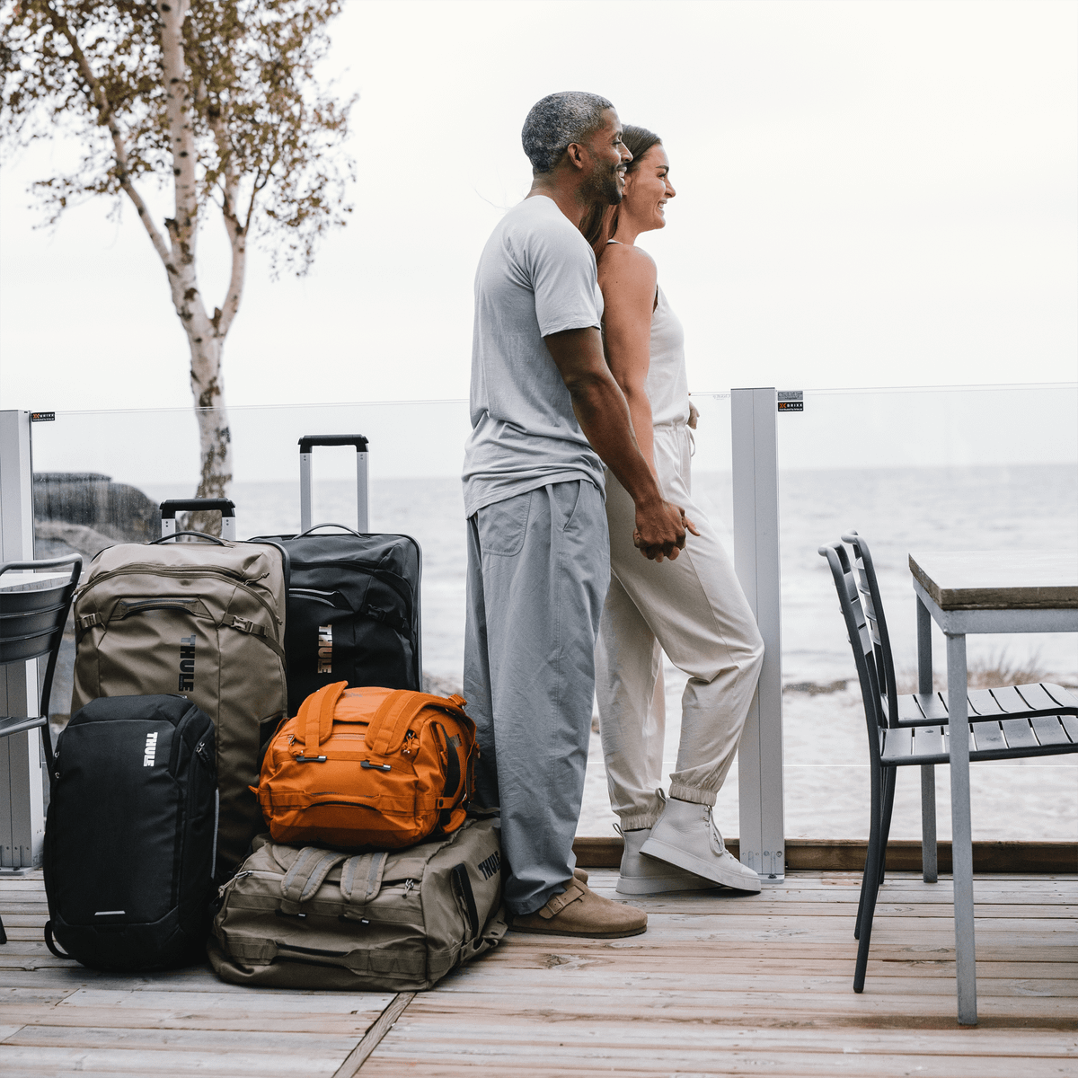A couple stand and look out at the ocean with a pile of Thule Chasm duffels and suitcases beside them.