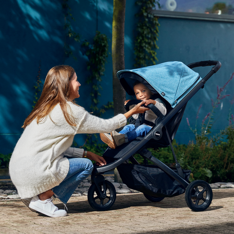 A woman kneels and tends to her toddler in a blue stroller who is holding the Thule Spring Bumper Bar.