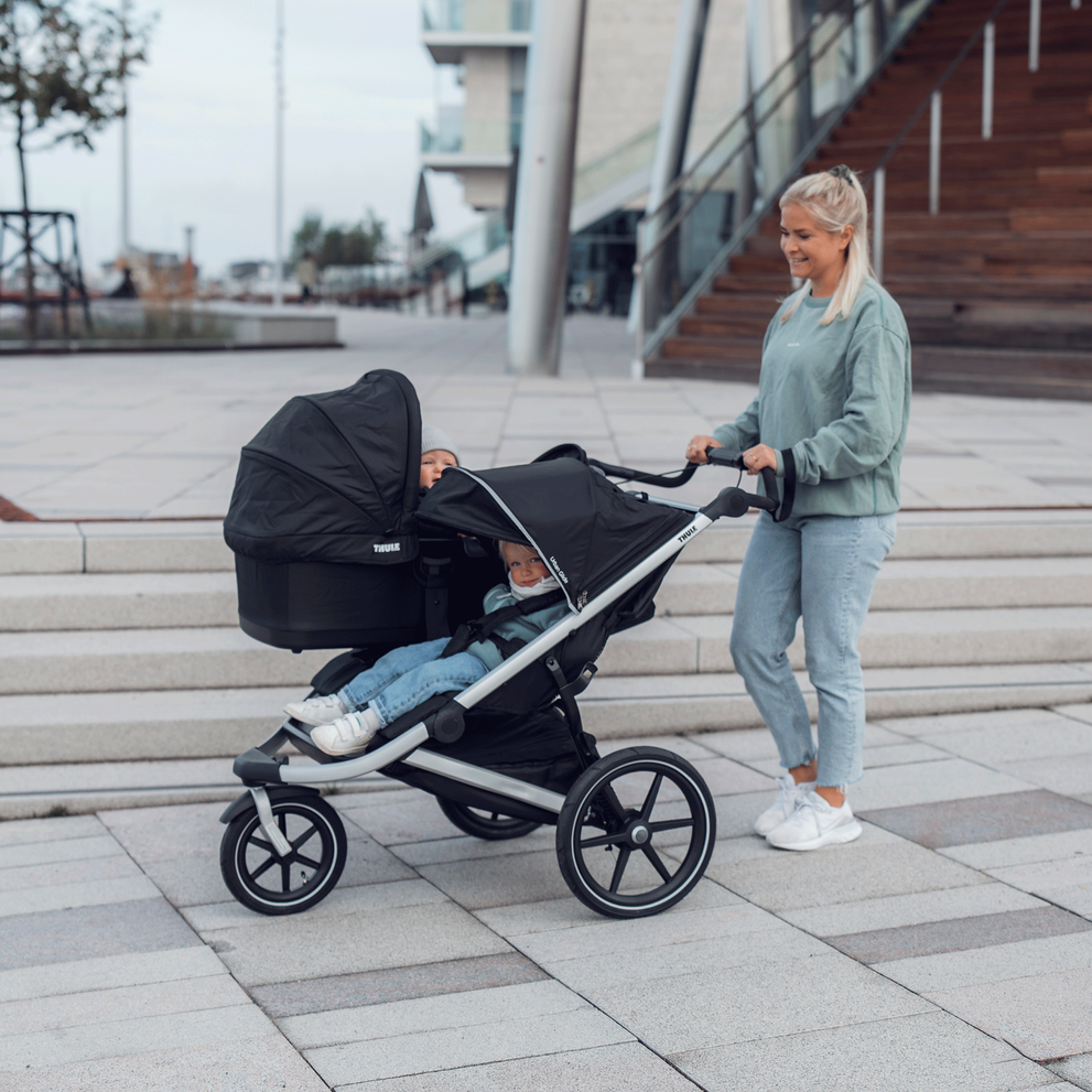A woman pushes her black Thule Urban Glide 2 double jogging stroller with a bassinet and toddler seat.