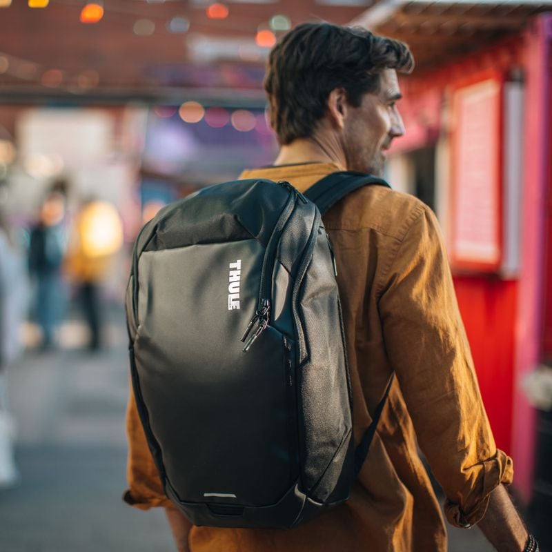 A man in an orange shirt is carrying a black Thule Chasm backpack.