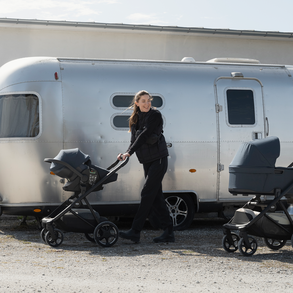 Adult pushing a stroller with a Thule Maple Connect infant car seat attached to it beside a camper trailer outdoors.