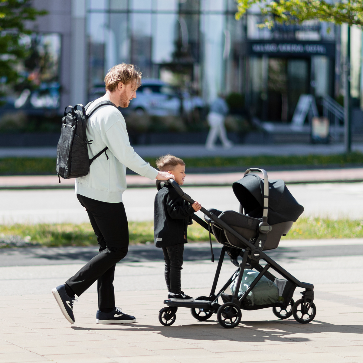 A parent walks with one kid on a rider board and the other in a car seat using the Thule Shine Car Seat Adapter.