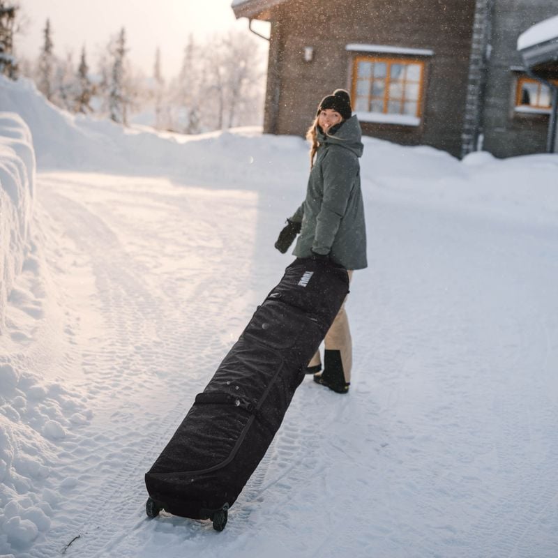 A woman stands in the snow outside a cottage wheeling her Thule RoundTrip travel snowboard bag.