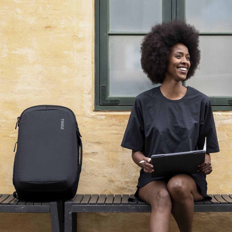 A close-up of  a woman sitting on a wooden bench with a tablet and a Thule Subterra travel backpack.