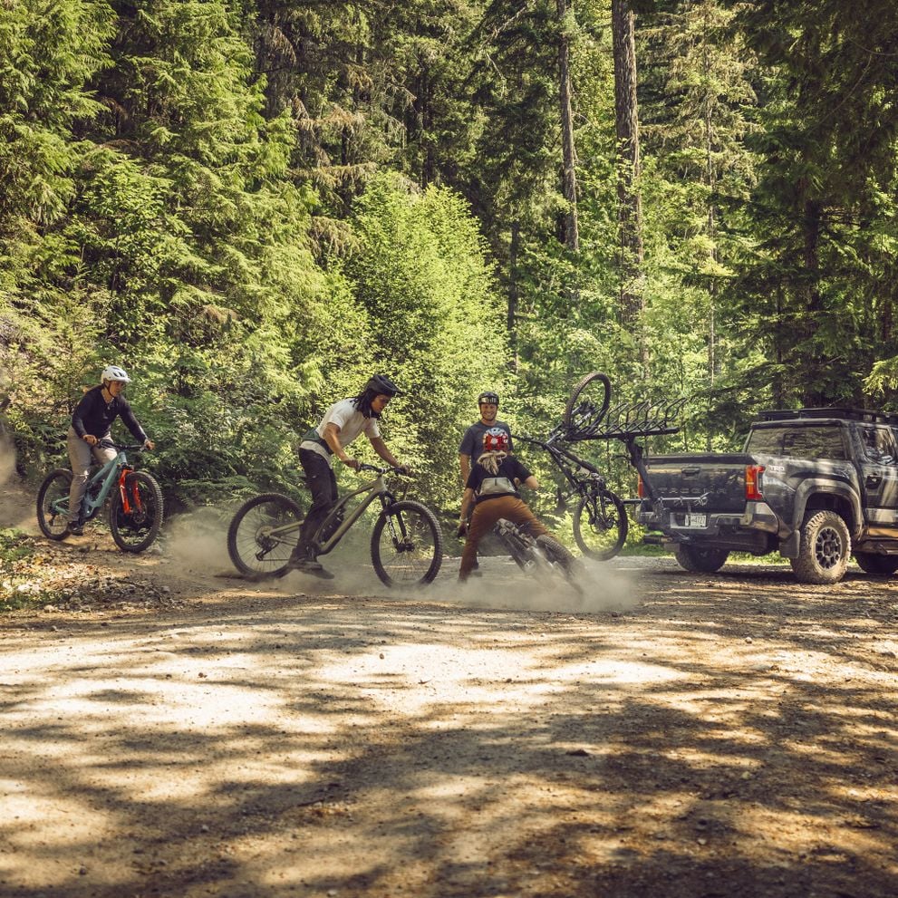 A pickup truck wiith the vertical Thule ReVert hitch bike rack is parked on a mountain road, while four persons are having fun riding around on their mountain bikes close to the pickup truck.