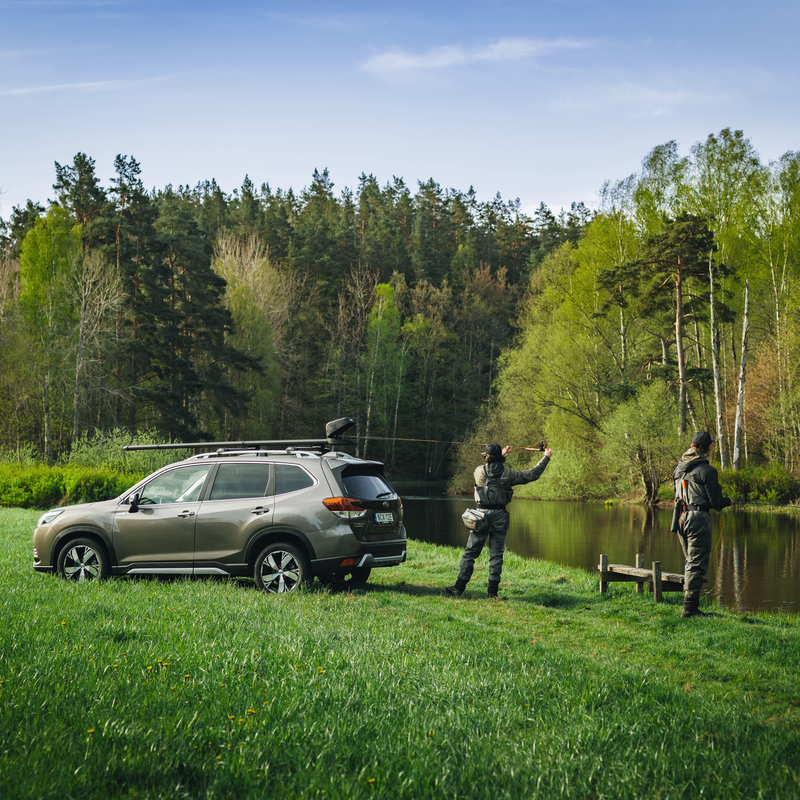Two men are fly fishing next to a river in a forest setting beside a car with a Thule RodVaule 2 fly rod carrier installed on the roof of the car.