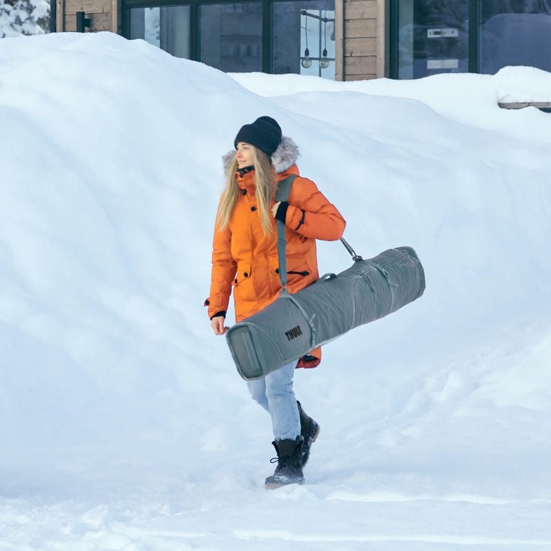 A woman in an oragnge ski jacket carries skis in a Thule RoundTrip ski bag, walking in a snowy setting.