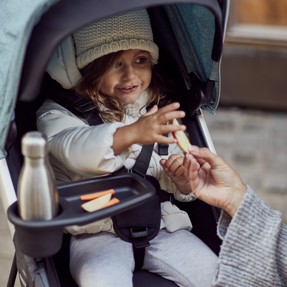 A toddler in a blue stroller has an apple and thermos on her Thule Spring Snack Tray.