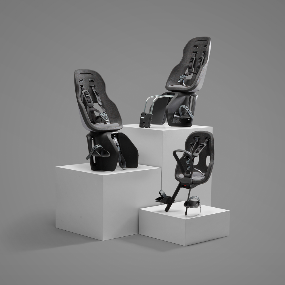 Three child bike seats are displayed at different heights on white pedestals against a grey background. Each seat features a sleek design with an adjustable safety harness and foot straps