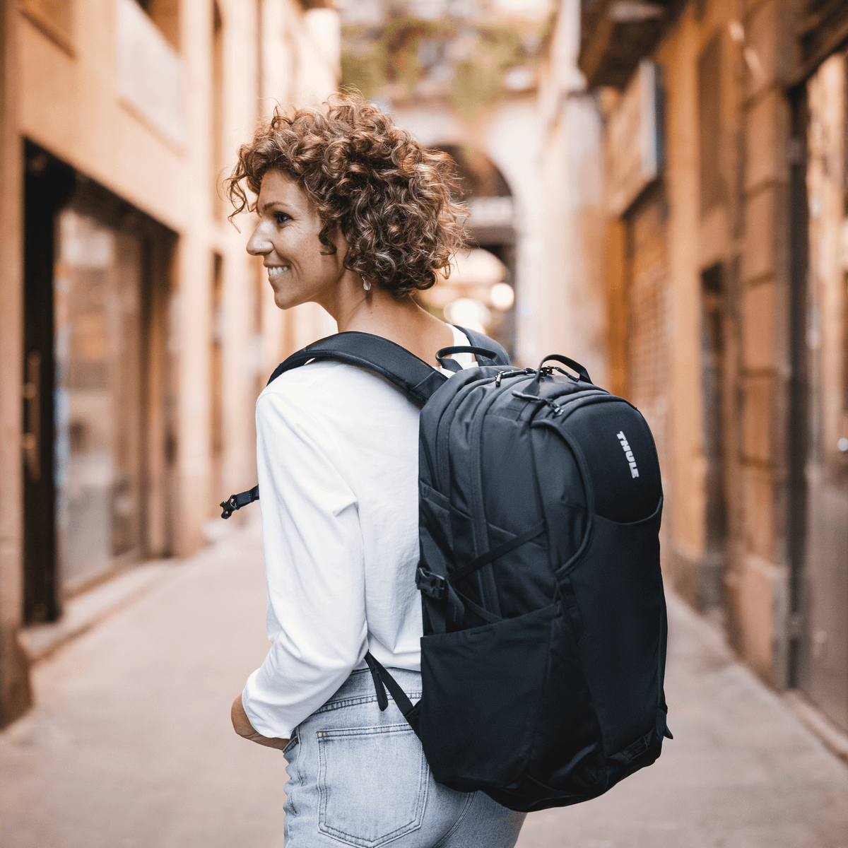 A woman walks down a sunny, narrow street with a black Thule Enroute backpack.