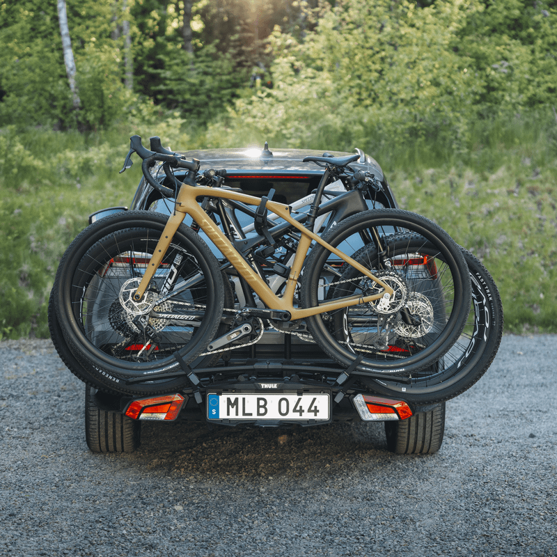 Three bicycles stand on a Thule EasyFold 3 bike carrier attached to the rear of a car parked in front of a forest landscape.