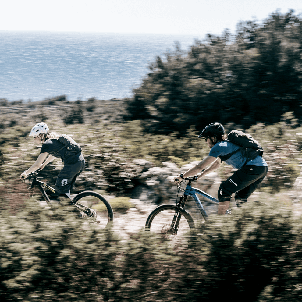 By the ocean, two people go mountain biking down  a mtb trail with a Thule Vital hydration pack.
