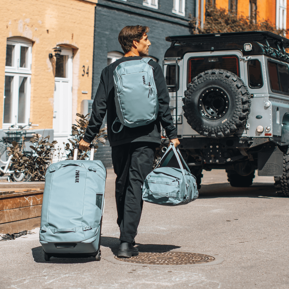 A man walks down a colorful street with blue Thule Chasm backpack, suitcase and duffel.