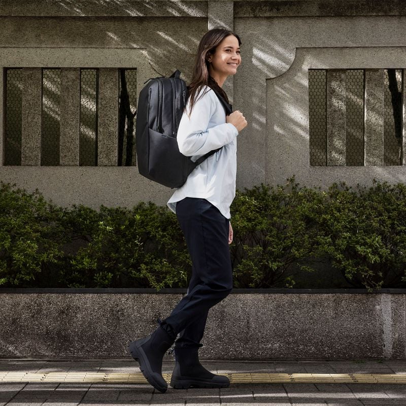 A woman walks down a sunny street while carrying a black Thule Subterra backpack.