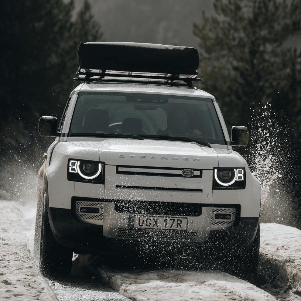 A car is driving on a snowy road with a roof top tent attached to the roof