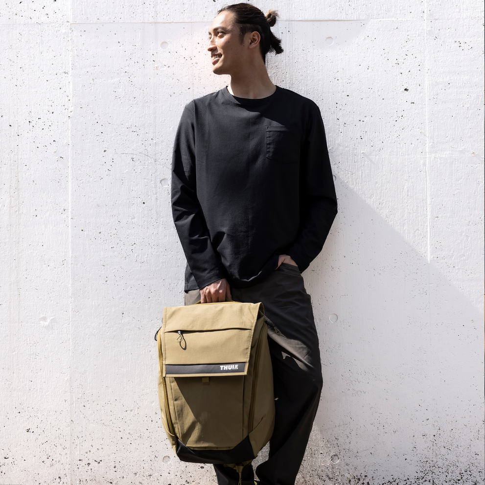 Man stands with back against white wall with Thule Paramount backpack 27L.