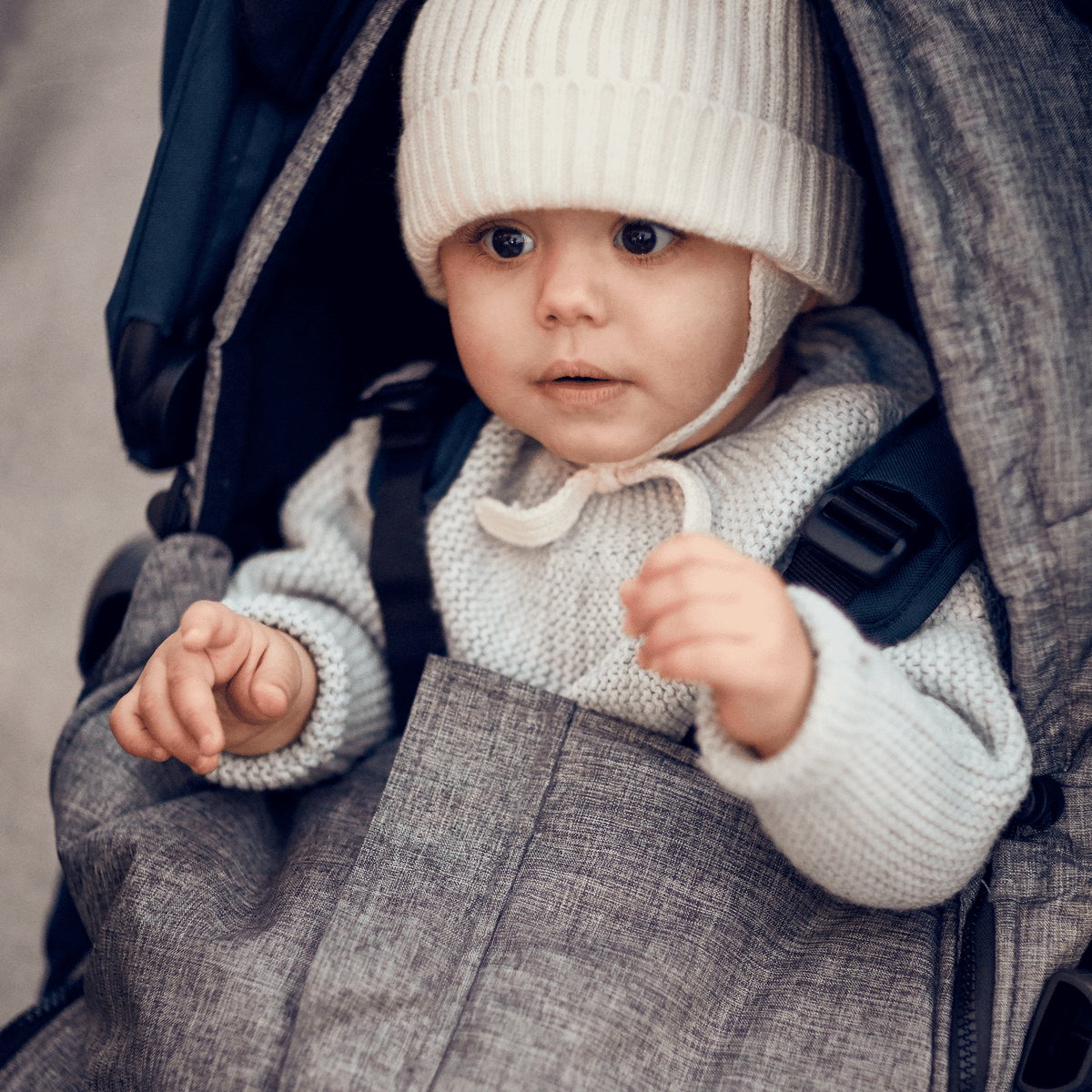 A baby with a fluffy hat sits in a stroller with a gray Thule Footmuff.