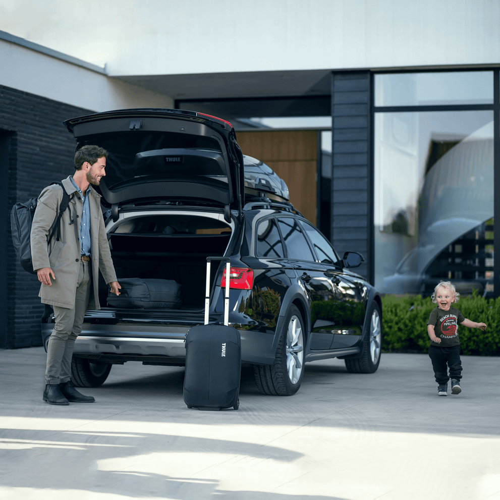 A man opens the trunk of his car, and a kid runs around as he takes out his Thule Subterra Carry-On suitcase.