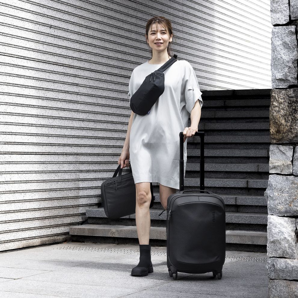 A woman walks down stairs with a black Thule Subterra suitcase, sling and briefcase.