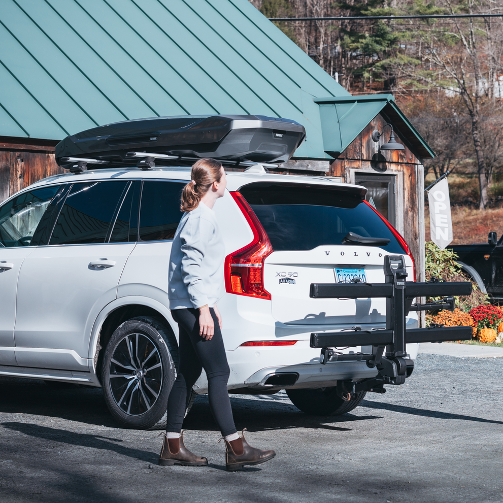 A woman stands next to a car with Thule Vero hitch bike rack tilted up and mounted on the back of a car parked in an outdoor setting.
