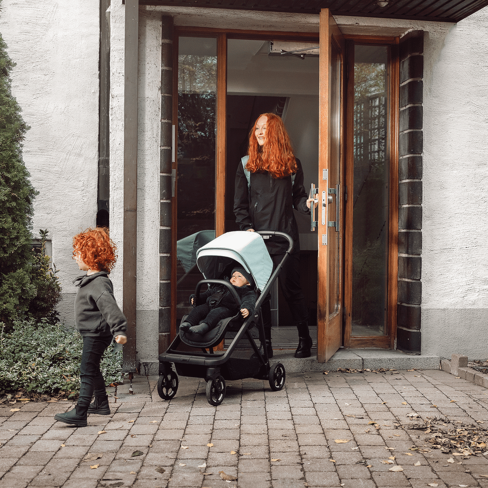 A woman exits her apartment building with a toddler running ahead and a baby in her Thule Shine stroller.