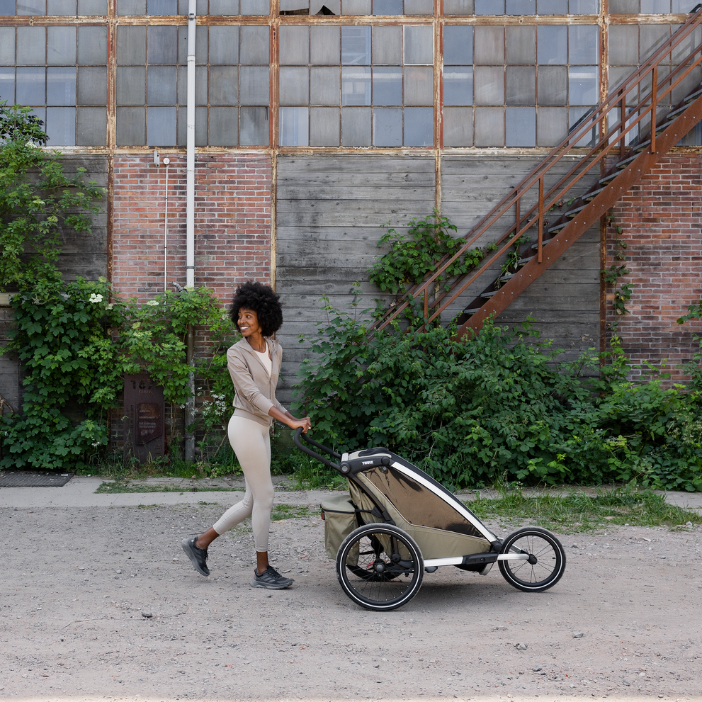 Parent walking outdoors while pushing a Thule Chariot Cross 2 multisport bike trailer and stroller, shown in an urban industrial area with greenery and brick walls in the background.