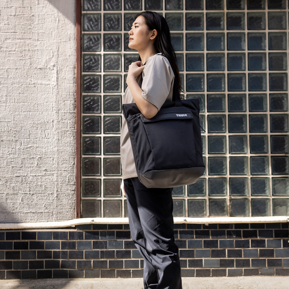 A woman stands with a glass wall holding a Thule Paramount tote bag.