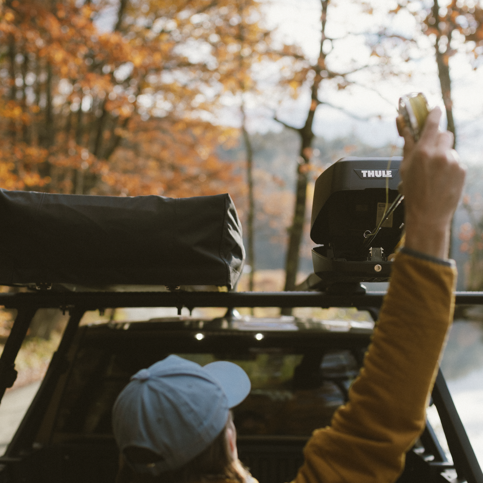 A man is feeding his fly rods into a Thule RodVault 2 fly rod carrier installed on the roof of a car in a forest setting.